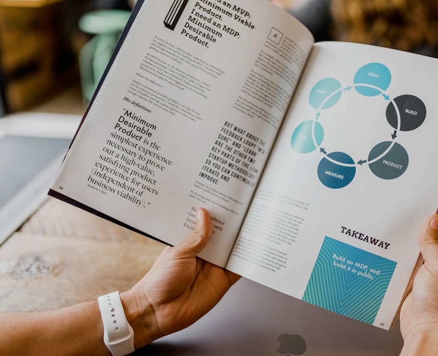 A person holding an open book displaying an article on the 'Minimum Desirable Product' concept, along with a circular flow diagram representing steps like 'Ideas', 'Build', 'Product' and 'Measure'. Text on the opposite page describes the Minimum Viable Product (MVP) strategy and key takeaways.