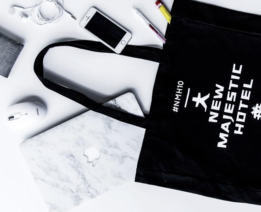 A flat lay of various objects on a white background, including a black tote bag with 'New Majestic Hotel' branding, a MacBook with a marble cover, a white wireless mouse, a smartphone, earbuds, and pens, presenting a neatly arranged and minimalist setup.