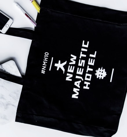 A flat lay of various objects on a white background, including a black tote bag with 'New Majestic Hotel' branding, a MacBook with a marble cover, a white wireless mouse, a smartphone, earbuds, and pens, presenting a neatly arranged and minimalist setup.