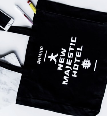 A flat lay of various objects on a white background, including a black tote bag with 'New Majestic Hotel' branding, a MacBook with a marble cover, a white wireless mouse, a smartphone, earbuds, and pens, presenting a neatly arranged and minimalist setup.