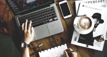 A workspace setup on a wooden table with a MacBook, iPhone, coffee cup with latte art, and a sketchpad where a hand is drawing the words 'Creativity is Contagious, Pass it On' in a decorative script. There's also a magazine visible with 'Yohji Yamamoto' on its cover. The scene is photographed from above in a lifestyle composition.