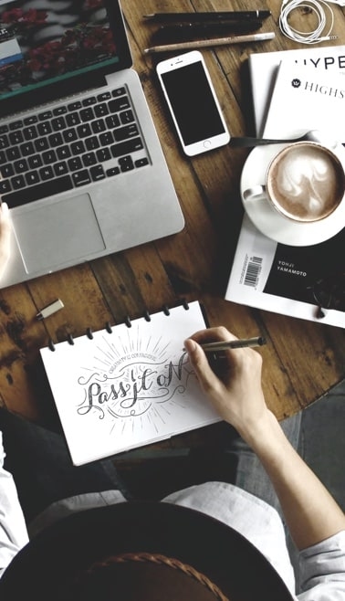 A workspace setup on a wooden table with a MacBook, iPhone, coffee cup with latte art, and a sketchpad where a hand is drawing the words 'Creativity is Contagious, Pass it On' in a decorative script. There's also a magazine visible with 'Yohji Yamamoto' on its cover. The scene is photographed from above in a lifestyle composition.