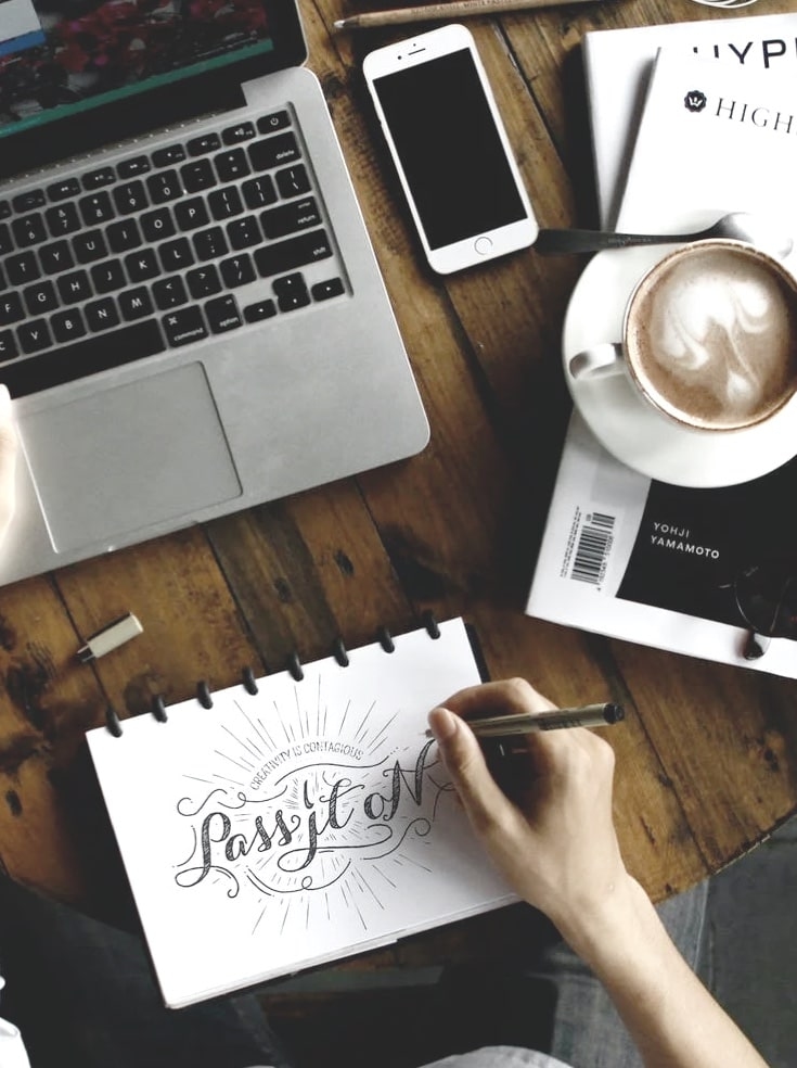 A workspace setup on a wooden table with a MacBook, iPhone, coffee cup with latte art, and a sketchpad where a hand is drawing the words 'Creativity is Contagious, Pass it On' in a decorative script. There's also a magazine visible with 'Yohji Yamamoto' on its cover. The scene is photographed from above in a lifestyle composition.